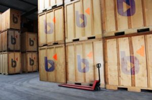 Boîtes et caisses en bois stockées dans un entrepôt, chariot élévateur devant, prêt à déménager où l’on peut se demander quand commencer à préparer son déménagement.