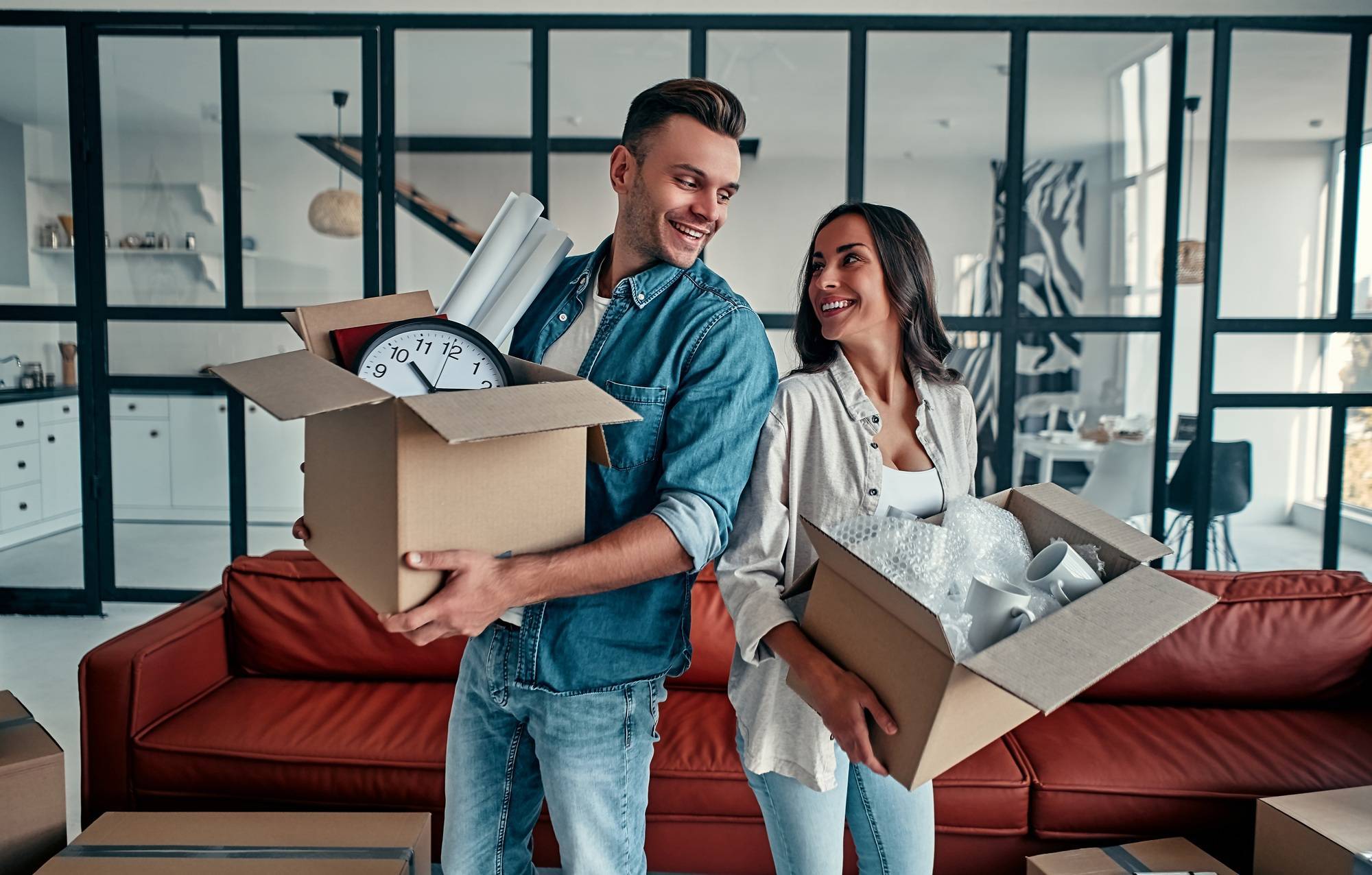 Couple souriant, portant des cartons d’emménagement remplis d’objets, dans leur salon moderne. Intégrer le mot-clé: “comment emballer ses affaires pour un déménagement” naturellement dans la description.