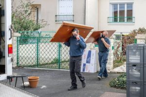 Deux déménageurs portent des meubles lourds en carton et planches, marchant devant une maison avec une clôture verte. Taille carton standard déménagement suggérée.