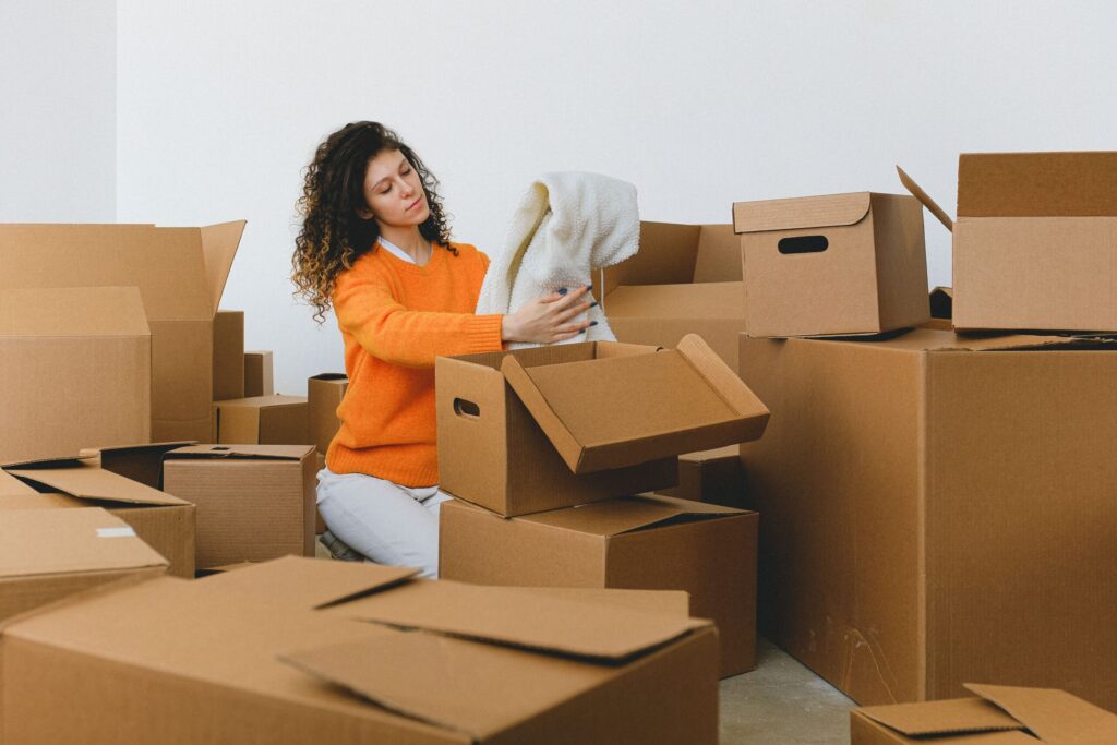 Femme en pull orange entourée de cartons, en train de préparer et fermer des boîtes lors d’un déménagement. Inclut le geste pour scotcher et sécuriser.
