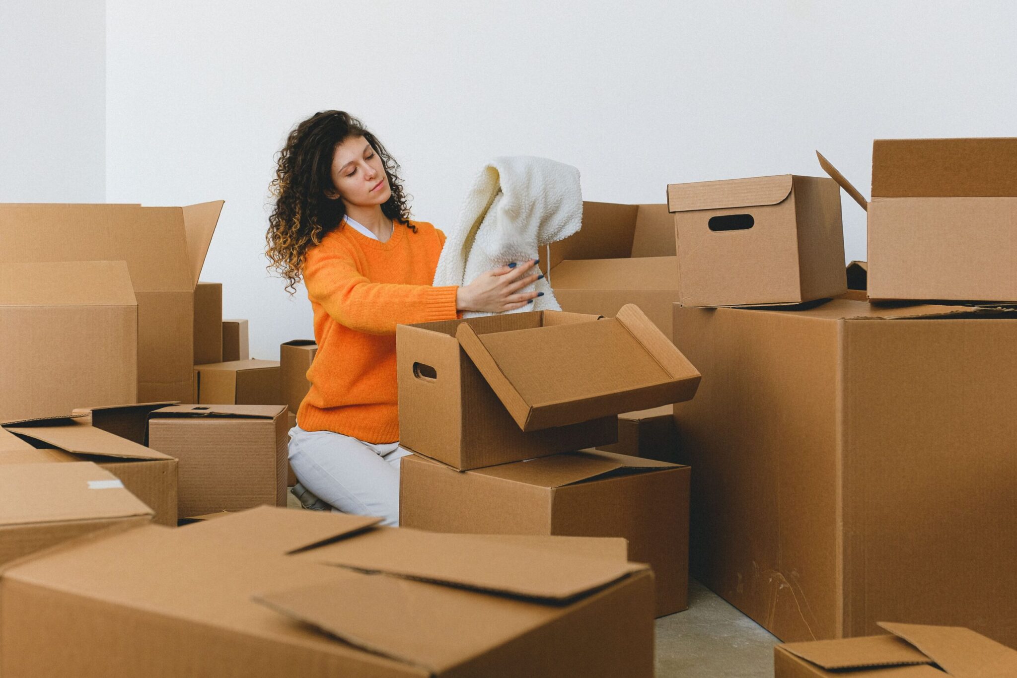 Femme en pull orange entourée de cartons, en train de préparer et fermer des boîtes lors d’un déménagement. Inclut le geste pour scotcher et sécuriser.