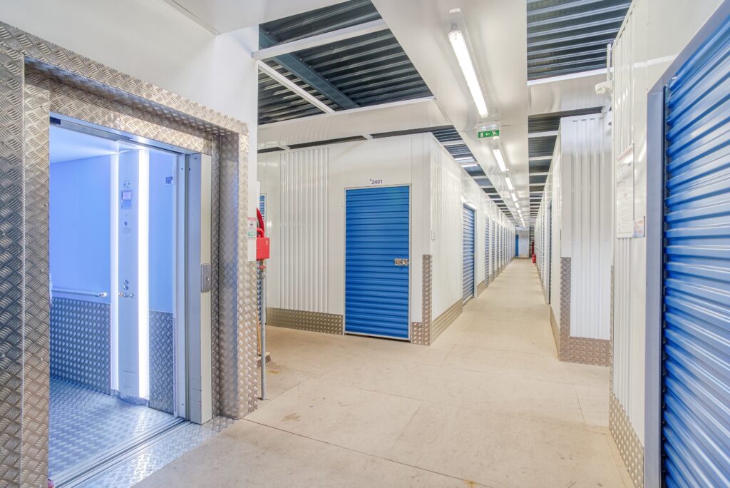 Couloir lumineux d’un centre de self-stockage avec des rangées de cabines à portes bleues et un ascenseur à gauche.