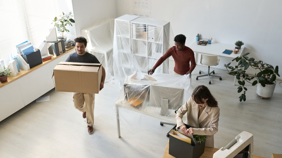 Trois personnes emballent et déplacent des cartons dans un bureau meublé, avec des meubles couverts et des objets de